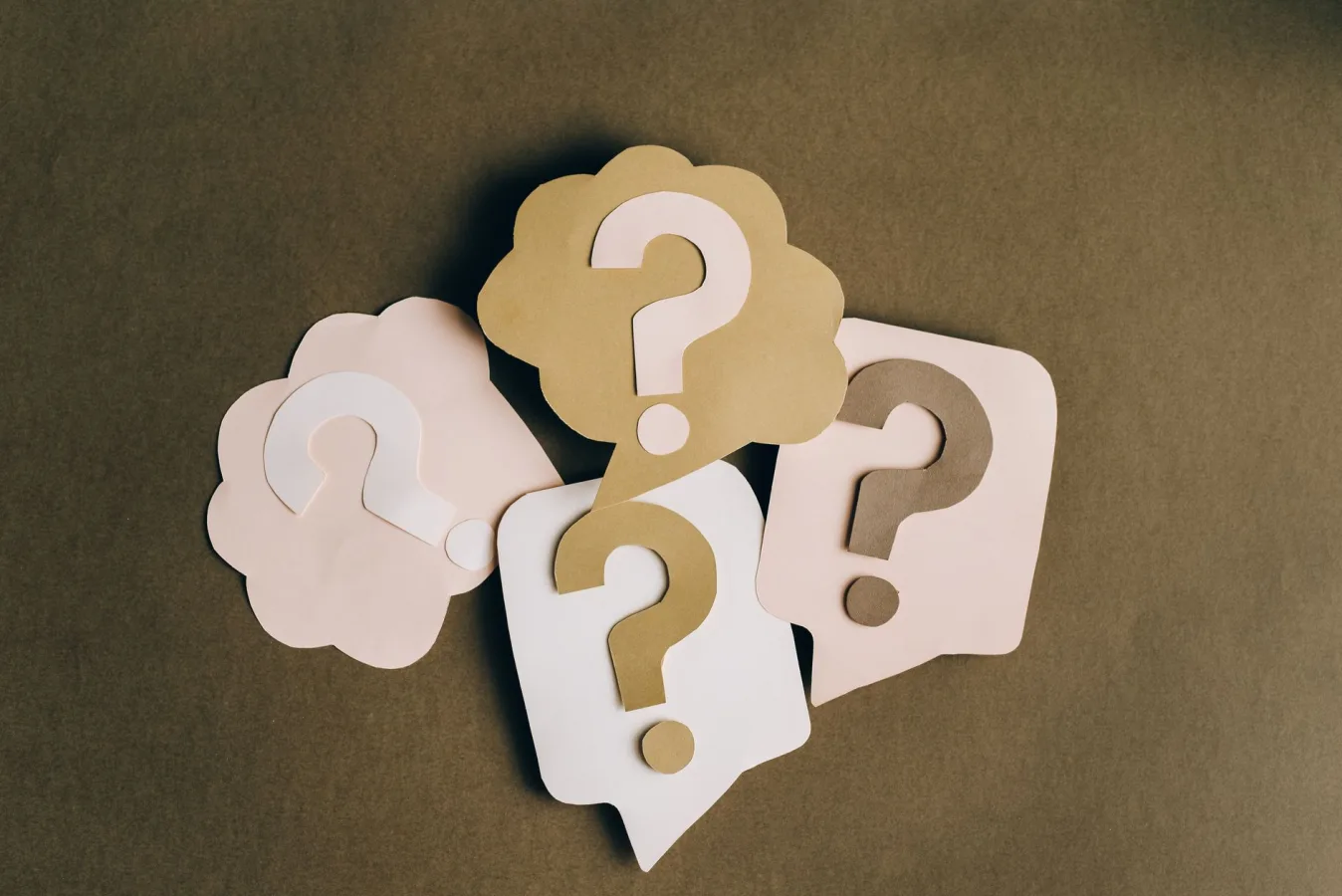 Four paper cutouts of question marks in beige and white on a brown surface, symbolizing in