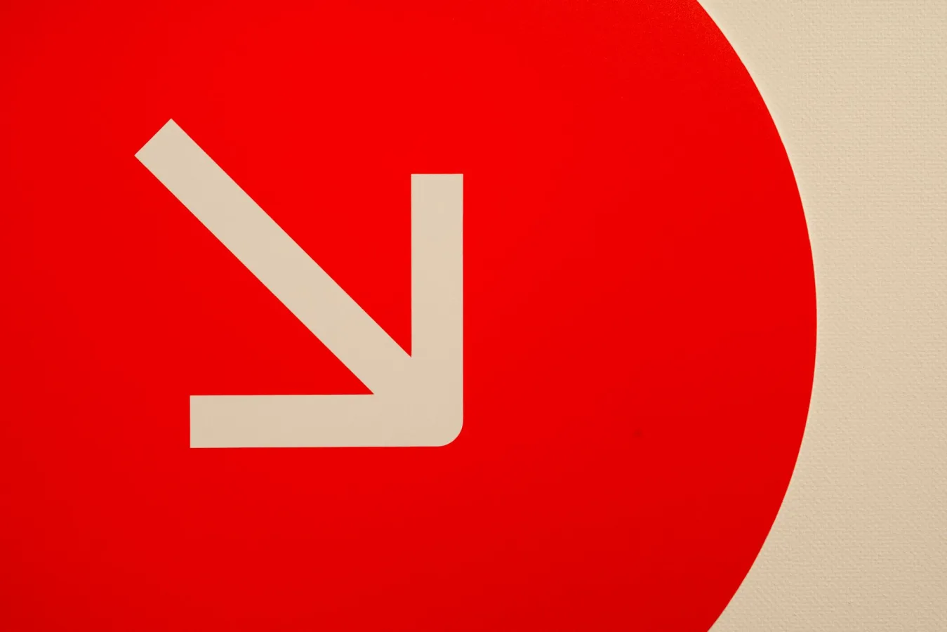 A minimalist image of a white downward arrow on a red round background, conveying directio