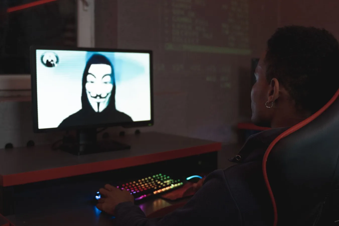 Person using computer in dark room viewing hacker wearing a mask on the screen, symbolizin