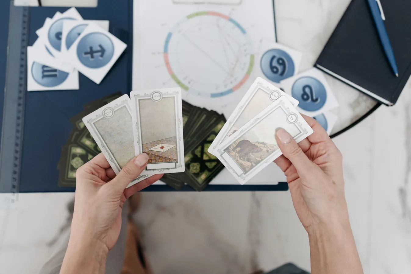 Hands holding tarot cards above an astrological chart with zodiac symbols scattered on the