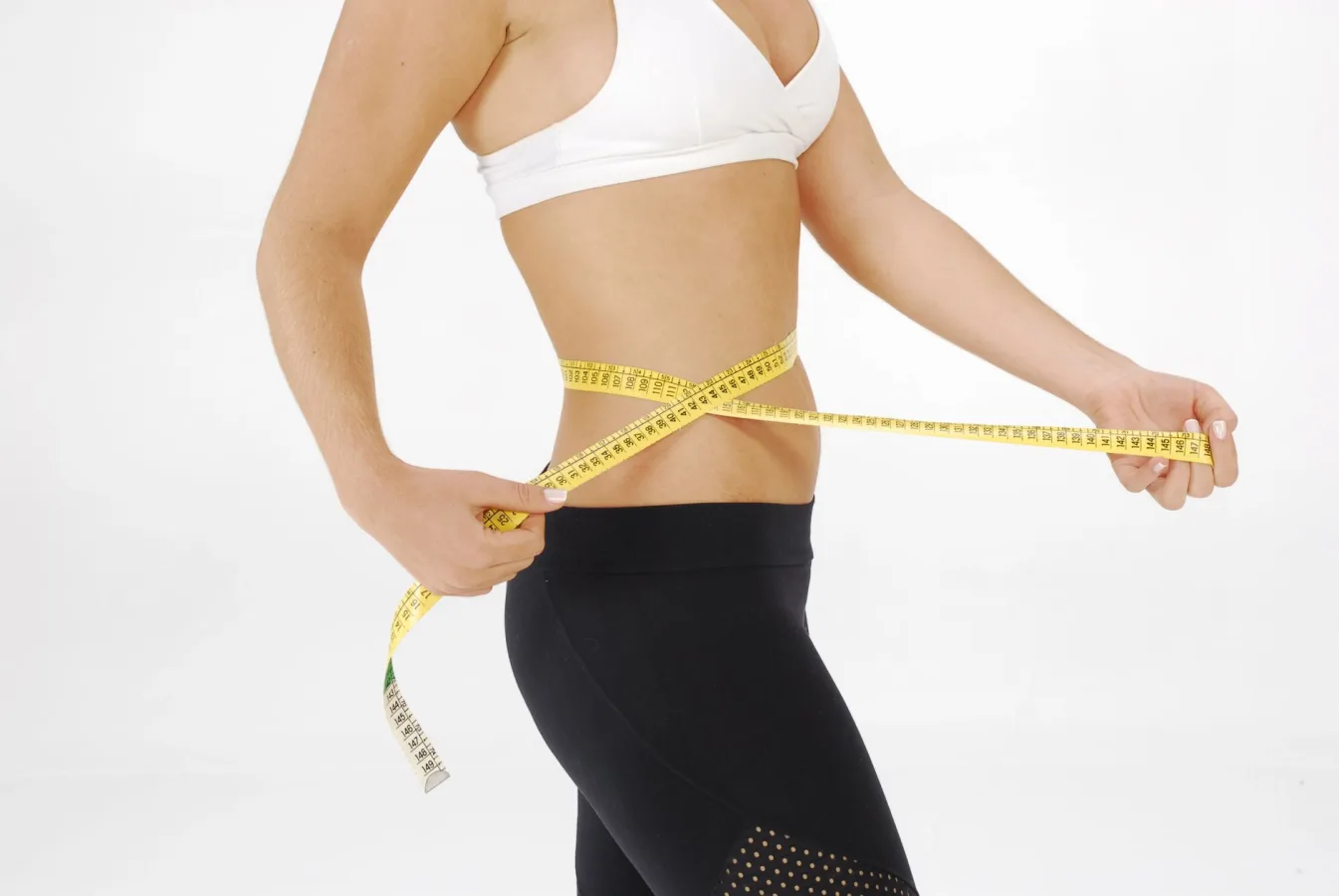 Fit woman in sportswear measuring her waist with a tape measure against a white background