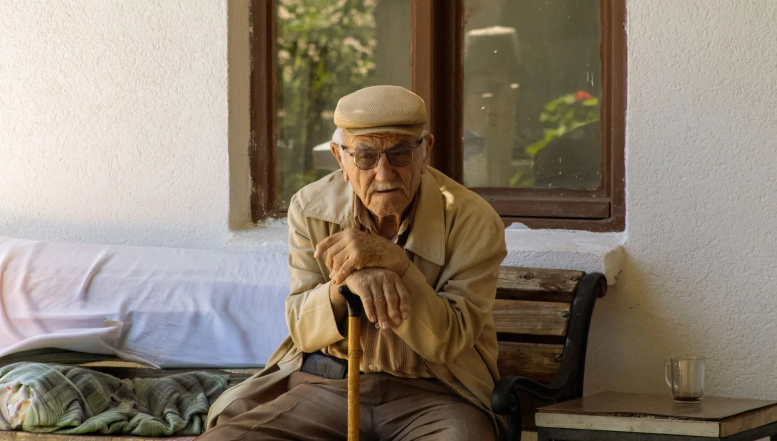 Senior man with glasses and a cap sitting outdoors on a bench, holding a cane.